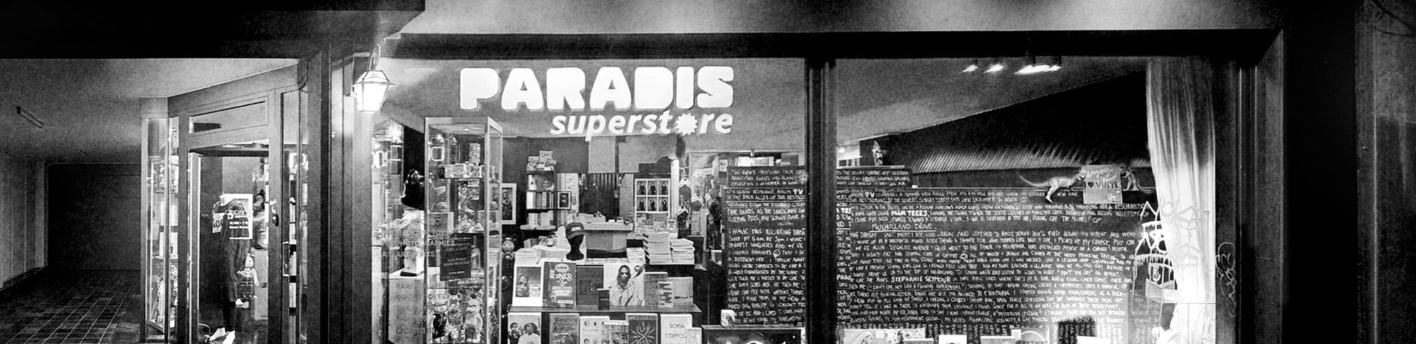 Black and white image of a store front with 'Paradis Superstore' sign and date display.